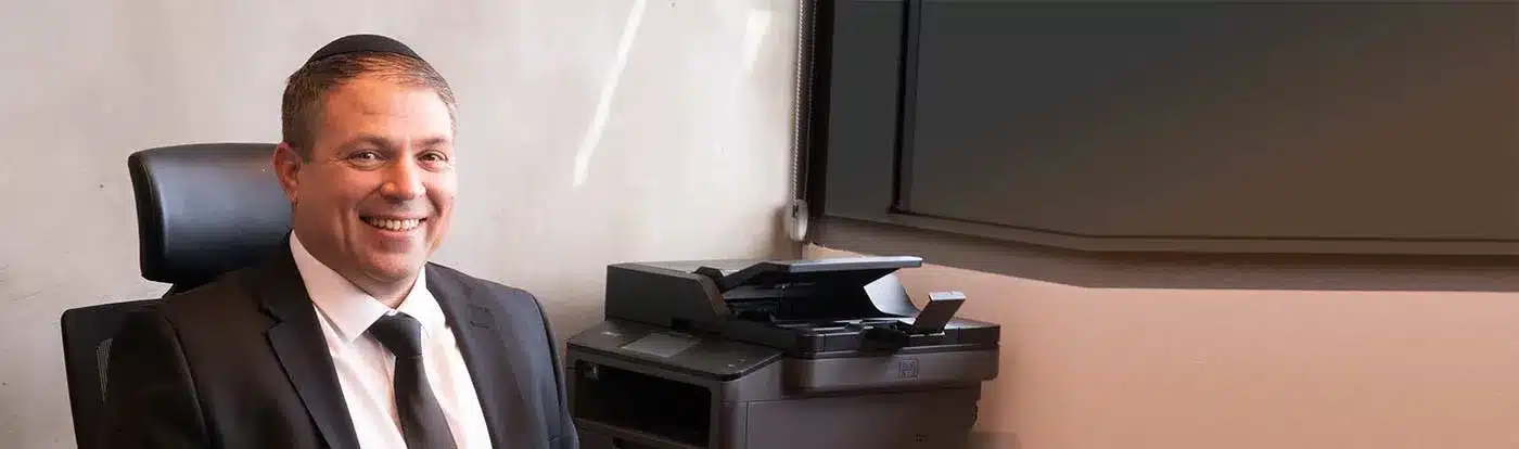 גבר בחליפה ועניבה יושב בכיסא משרדי ליד מדפסת, מחייך למצלמה.
